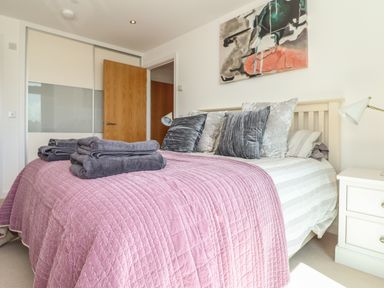 A bedroom with a bed covered by a pink quilt and stacked gray towels at 8 Cribbar in Newquay