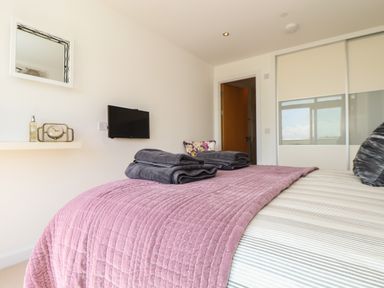 A bedroom with bed linens and folded towels on the bed a wall-mounted television a shelf with a clock and a bottle a door and a window closet with sliding doors at 8 Cribbar in Newquay