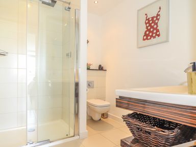 A bathroom with a glass shower enclosure a wall-mounted toilet a wooden vanity with a basket and a framed picture on the wall at 8 Cribbar in Newquay