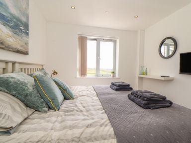 A bedroom with a bed with pillows and folded towels on it a window with partial blinds a wall-mounted TV and a round mirror at 8 Cribbar in Newquay