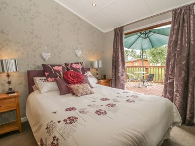 A bedroom with a bed featuring floral bedding and pillows bedside tables with lamps and an open door to a patio with outdoor furniture and an umbrella at Bulmer Farm Lodge in Malton