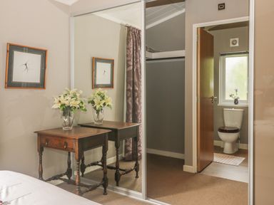 A bedroom corner with a wooden side table holding a vase of white flowers a large mirrored closet door and a view into a bathroom with a toilet and window at Bulmer Farm Lodge in Malton