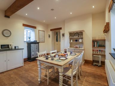A dining room with a wooden table set for breakfast with plates mugs and pastries surrounded by chairs and cupboards at Woodlands Cottage in Docklow near Leominster