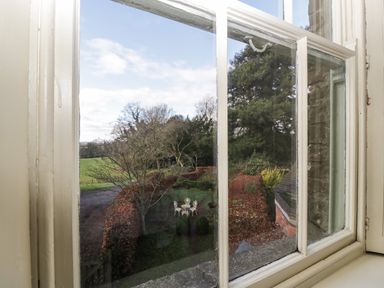 A view through a window showing a garden with trees hedges and a white metal table and chairs at Woodlands Cottage in Docklow near Leominster