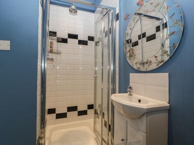 A bathroom with a shower area with white and black tiles a white sink with a cabinet and a round mirror on a blue wall at Woodlands Cottage in Docklow near Leominster