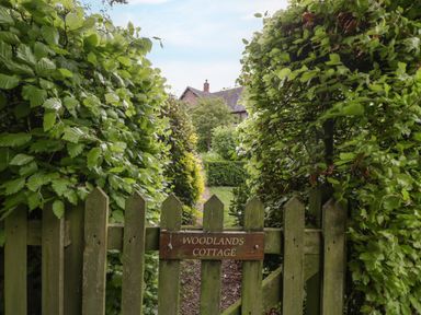 A wooden garden gate with a woodlands cottage sign surrounded by green bushes at Woodlands Cottage Docklow near Leominster