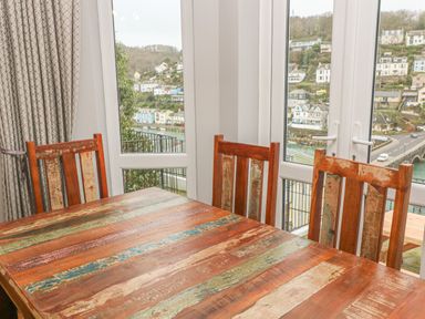 A dining table with wooden chairs by windows showing a hillside with houses and a river at Elmbank in Looe