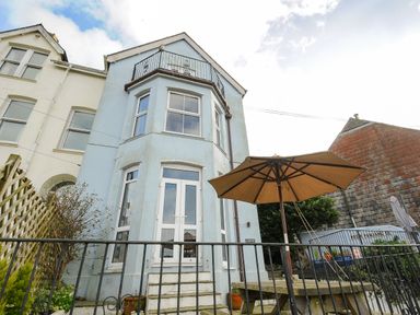 The rear exterior of a light blue house with upper balcony and patio table with umbrella at Elmbank in Looe