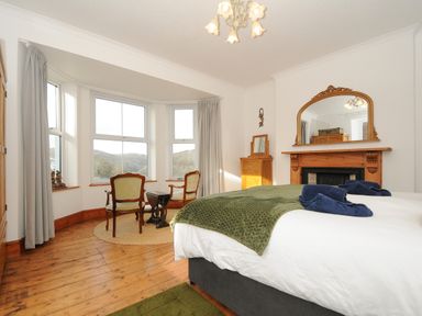 A bedroom with a bed near a fireplace and two chairs by bay windows at Elmbank in Looe