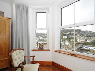 A corner of a room with a wooden chair, an armoire, statues on a window sill and a view of a town and river through the windows at Elmbank in Looe