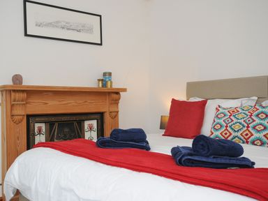 A bedroom with a bed covered in white bedding and red and patterned cushions near a wooden fireplace with decorative tiles at Elmbank in Looe