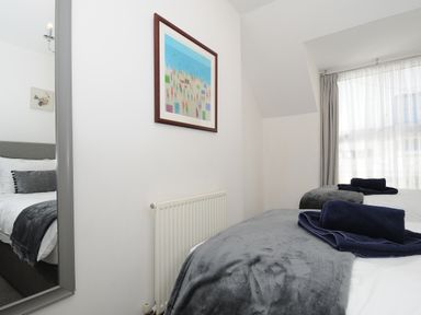 A bedroom with a bed covered with gray blanket and navy towels a radiator a framed picture on the wall and sheer curtains at Elmbank in Looe