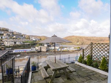 An outdoor patio with a wooden picnic table and umbrella overlooking a river and hillside houses at Elmbank in Looe