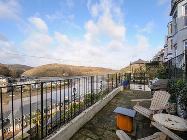 A balcony with wooden chairs, a table, a barbecue grill, and a railing overlooking a river and hillside at Elmbank in Looe