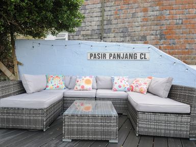 An outdoor corner sofa with gray cushions and colorful floral pillows on a wooden deck against a light blue wall with a street sign reading Pasir Panjang Cl