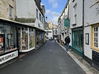 A narrow street with shops and signs including Pepe's Tex-Mex Kitchen and a vape supplies shop with people walking and standing at Elmbank in Looe