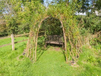 A garden bench inside a wicker arch surrounded by grass and trees at Little Freestone in Carew