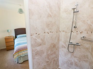 A shower area with beige tiles and a silver showerhead next to a bedroom with a striped bedspread and wooden bedside table at Little Freestone in Carew