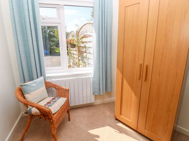 A small room with a wicker chair with cushions near a window and a wooden wardrobe at Little Freestone in Carew