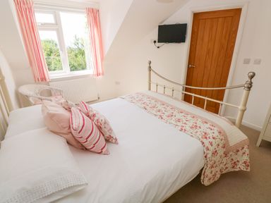 A bedroom with a bed with floral and striped pillows next to a window with pink curtains and a wicker chair at Little Freestone in Carew
