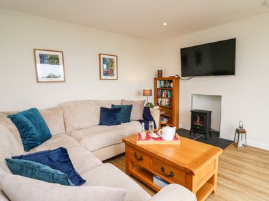 A living room with a beige sectional sofa with blue and gray cushions a wooden coffee table a bookshelf a floor lamp a mounted flat screen TV and an electric stove at Gwynant in Pontrhydfendigaid