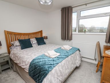A bedroom with a wooden bed frame bedside tables with lamps a window with brown curtains and a desk with a chair at Gwynant in Pontrhydfendigaid