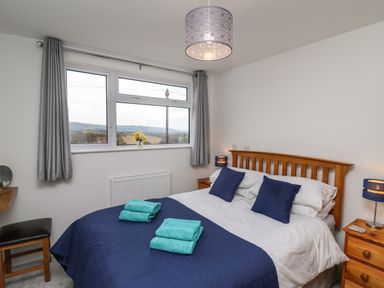 A bedroom with a double bed with blue bedding and pillows two bedside tables with lamps a window with gray curtains and a stool at Gwynant in Pontrhydfendigaid