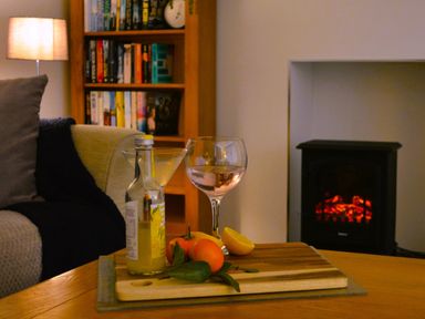 A living room with a wooden table holding a bottle, two glasses, fruits, a couch with cushions, a lamp, a bookshelf, and a fireplace at Gwynant in Pontrhydfendigaid