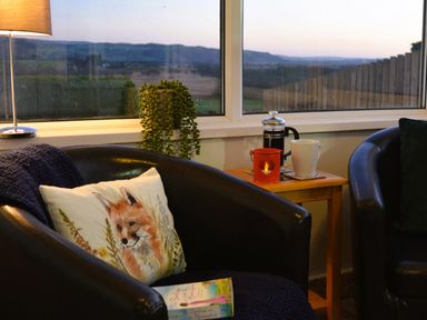 Two black chairs with cushions beside a wooden side table holding a coffee press a mug and a candle near a window with a landscape view at Gwynant in Pontrhydfendigaid