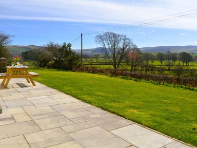 A patio with a wooden picnic table and a grassy yard with trees and hills in the background at Gwynant in Pontrhydfendigaid