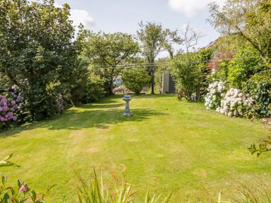 A garden with a grassy lawn surrounded by trees shrubs and flowers with a birdbath in the center at Glan Gors Llanerchymedd