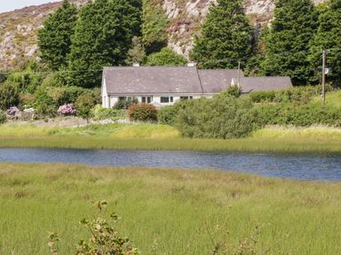 A house with a grey roof surrounded by bushes and trees near a body of water at Glan Gors in Llanerchymedd