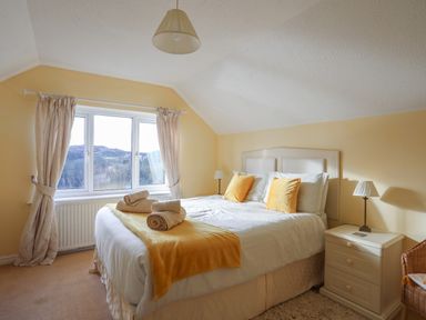 A bedroom with a double bed yellow pillows and throw bedside tables with lamps and a window with curtains at Glan Gors in Llanerchymedd