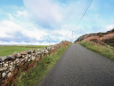 A narrow paved road with stone wall on left and grassy slopes on right in countryside at Glan Gors in Llanerchymedd