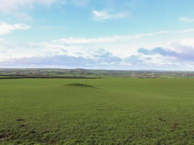 A green grassy field with a small mound and a pole in the center with distant hills and power lines at Glan Gors in Llanerchymedd