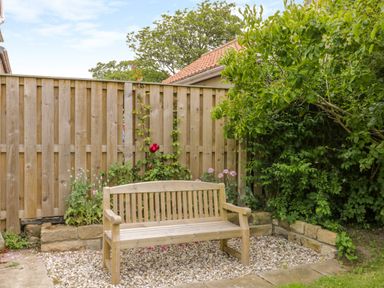 A wooden bench on a gravel area next to a wooden fence with plants and bushes at Stakesby House Apartment 1 in Whitby