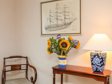 A corner with a wooden chair a wooden table with a blue floral lamp and a vase of sunflowers below a framed ship drawing at The Pink House in Rhosneigr