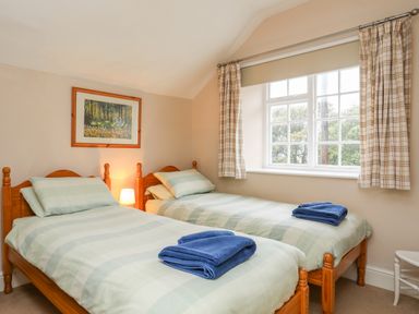 A bedroom with two single beds each with blue towels on top a window with checkered curtains a framed picture and a bedside lamp at The Pink House in Rhosneigr