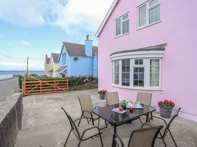 Outdoor patio area with table and chairs beside a pink house near a seaside road at The Pink House in Rhosneigr