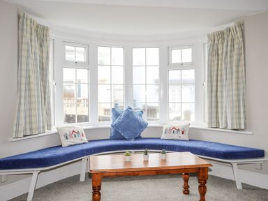 A curved window seat with blue cushions and pillows in front of a bay window with checkered curtains and a wooden coffee table with small plants at The Pink House in Rhosneigr