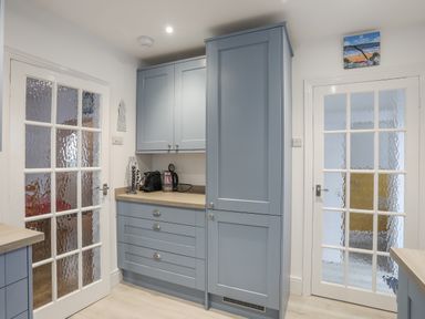 A kitchen area with blue cabinets wooden countertops a kettle a coffee machine and two glass paneled doors at The Pink House in Rhosneigr