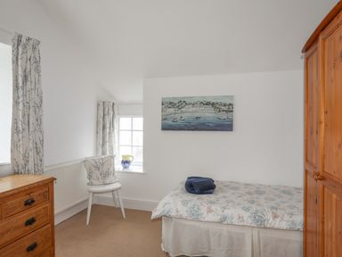 A bedroom with a single bed a wooden wardrobe a chest of drawers a chair with a cushion and a window with patterned curtains at The Pink House in Rhosneigr