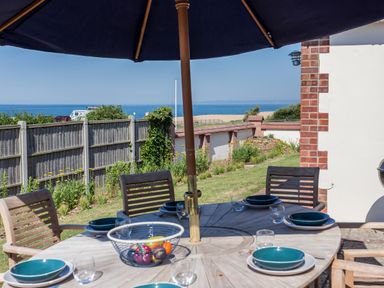 An outdoor dining table with six place settings under a large umbrella near a garden with a view of the sea at Beach Haven in West Bexington