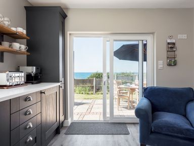 A kitchen area with dark cabinets and a coffee machine next to a sliding glass door opening to a patio with wooden chairs and an umbrella at Beach Haven in West Bexington