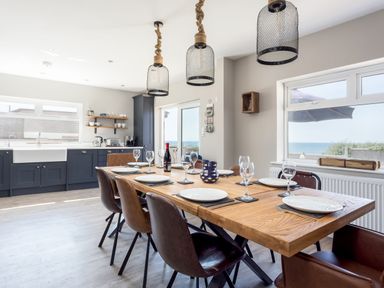 A dining area with a wooden table set with plates and glasses next to a kitchen with dark cabinets and a window showing the sea at Beach Haven in West Bexington