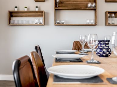 A dining table set with white plates wine glasses and a blue vase with fish pattern in a room with wooden shelves holding glasses at Beach Haven in West Bexington