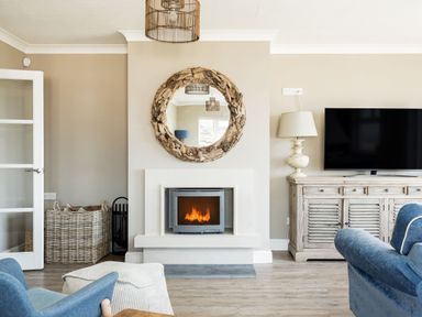 A living room with a wood burning stove fireplace mirror above basket lamp and television on wooden cabinet at Beach Haven in West Bexington