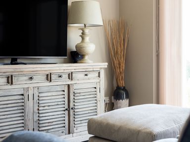 A wooden cabinet with a flat screen tv and a table lamp next to a vase with decorative sticks and an ottoman in a living room at Beach Haven in West Bexington