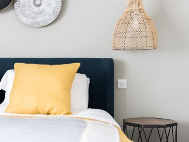 A bed with a yellow pillow white sheets and a blue headboard with a wooden side table a wicker lampshade and wall decorations at Beach Haven in West Bexington
