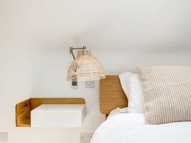 A bed with white bedding and a striped cushion next to a wooden floating nightstand and a wall lamp at Beach Haven in West Bexington